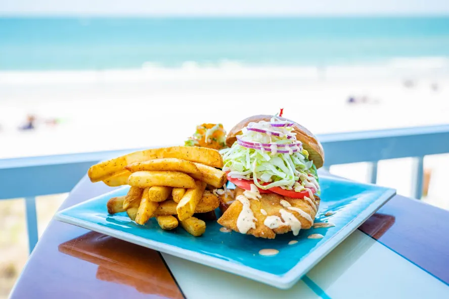 a fish sandwich and fries on a table that looks like a surfboard at Caddy's Madeira Beach, with a view of the beach in the distance