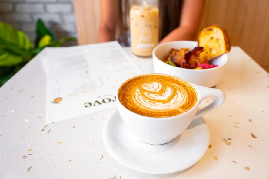 a hot latte with a pretty design next to an iced coffee and a breakfast bowl on a table at Grove Surf Cafe