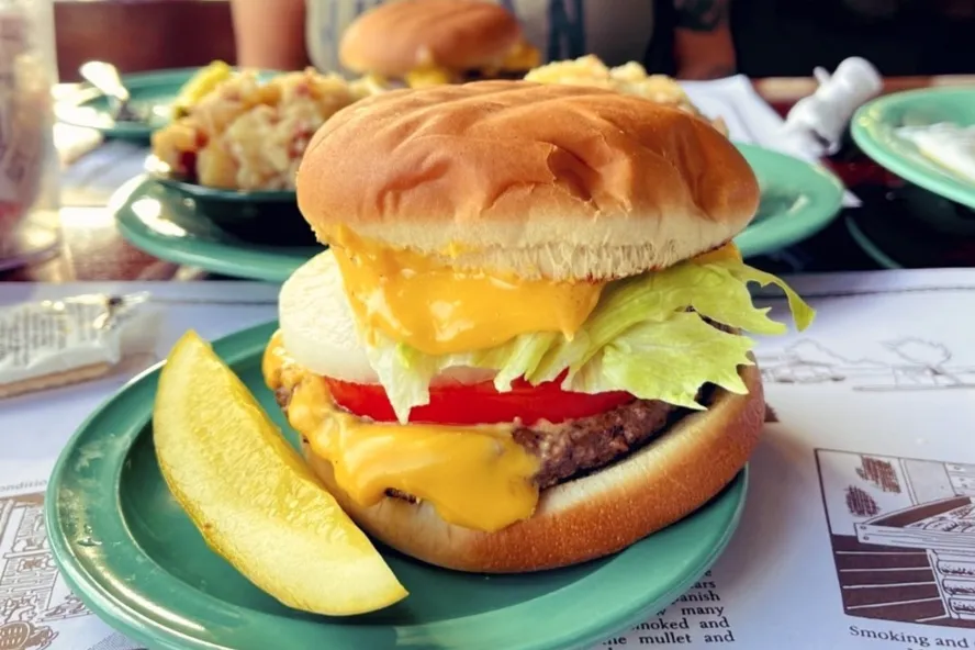 a cheeseburger with melted American cheese, onion, tomato and lettuce, next to a pickle wedge at Ted Peters Famous Smoked Fish