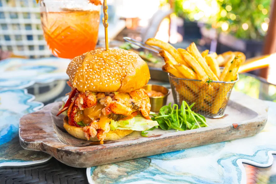 a wooden tray with a burger topped with butter-poached lobster on a sesame seed bun next to a basket of crispy fries at The Helm Seafood Parlour