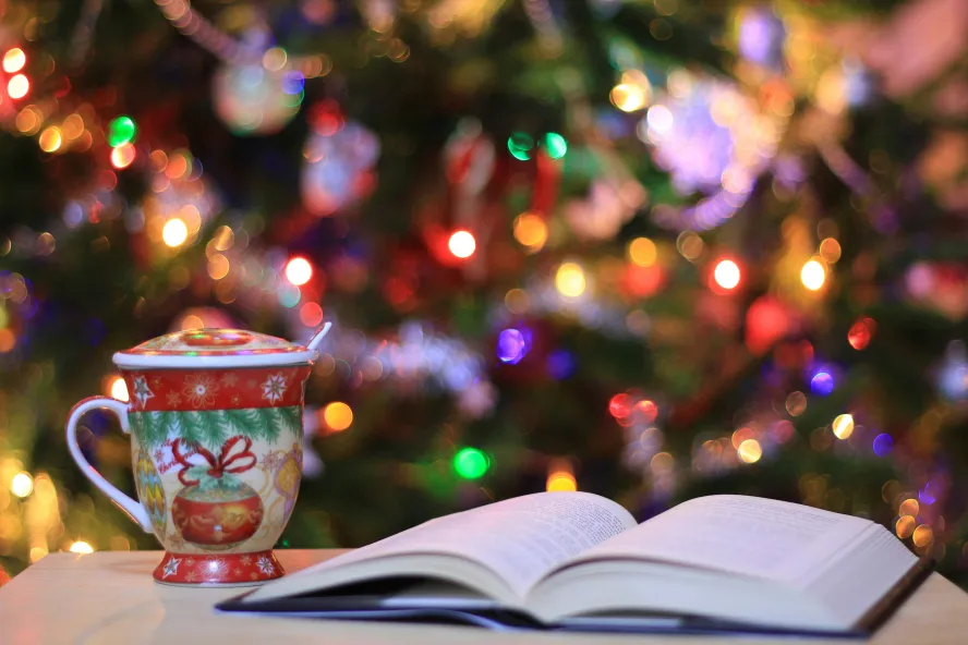 An open book on a counter with a tea mug and a Christmas tree lit up in the background.