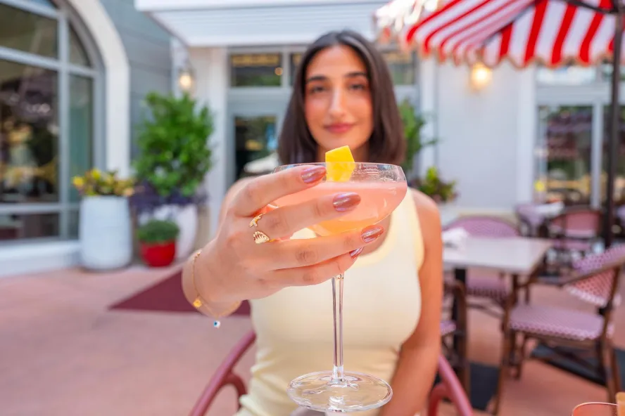 a woman holding out a cocktail toward the camera; she is sitting an an outdoor table near a striped umbrella at Juno & The Peacock