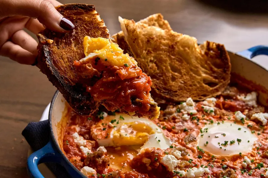 a person's hand dipping toasted bread into a large pan of Shakshouka during brunch at Elliott Aster at The Vinoy Hotel