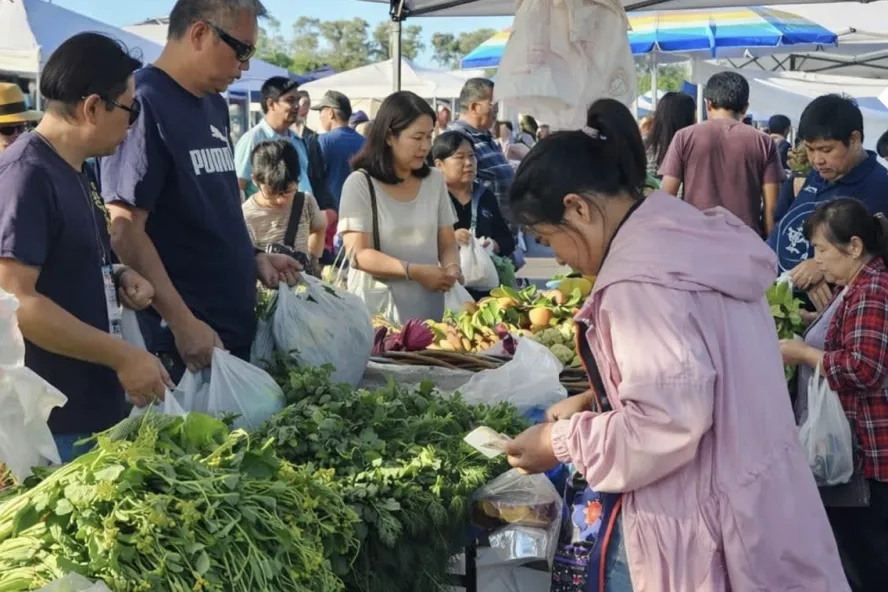 People shopping for fresh produce under a white vendor tent at an outdoor local farmers market.