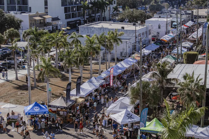 Aerial view of Mezzo Market with rows of vendor tents and crowds of shoppers filling a palm-lined street on a sunny day.