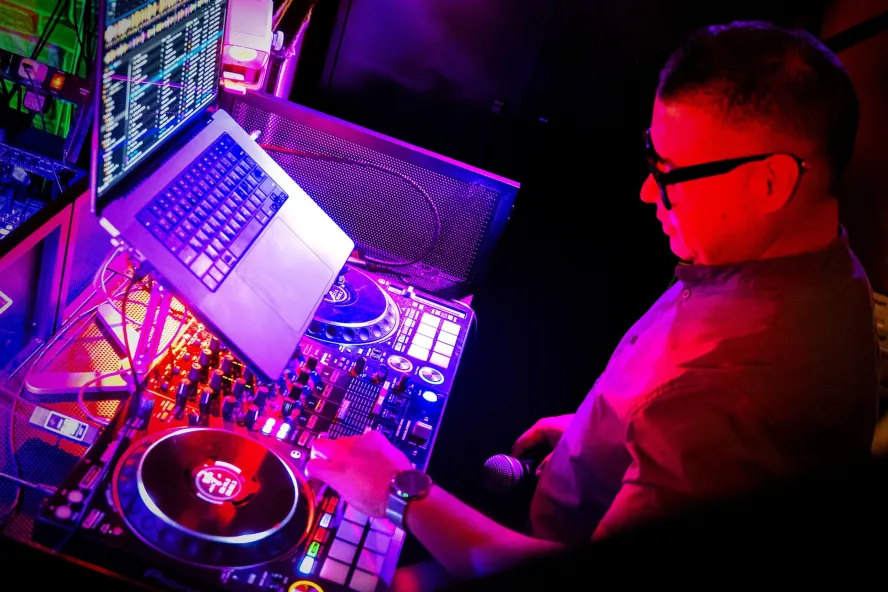 A DJ operates a mixing console and laptop beneath vibrant pink and purple lights at a nighttime dance event.