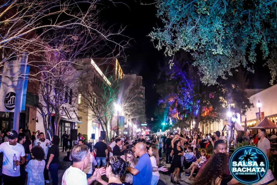 A lively crowd dances along a tree-lined street illuminated by colorful lights during Salsa & Bachata at The District in downtown St. Pete.