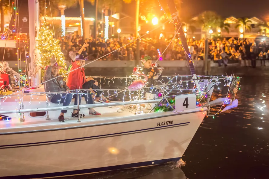 People waving on a sailboat wrapped in Christmas lights
