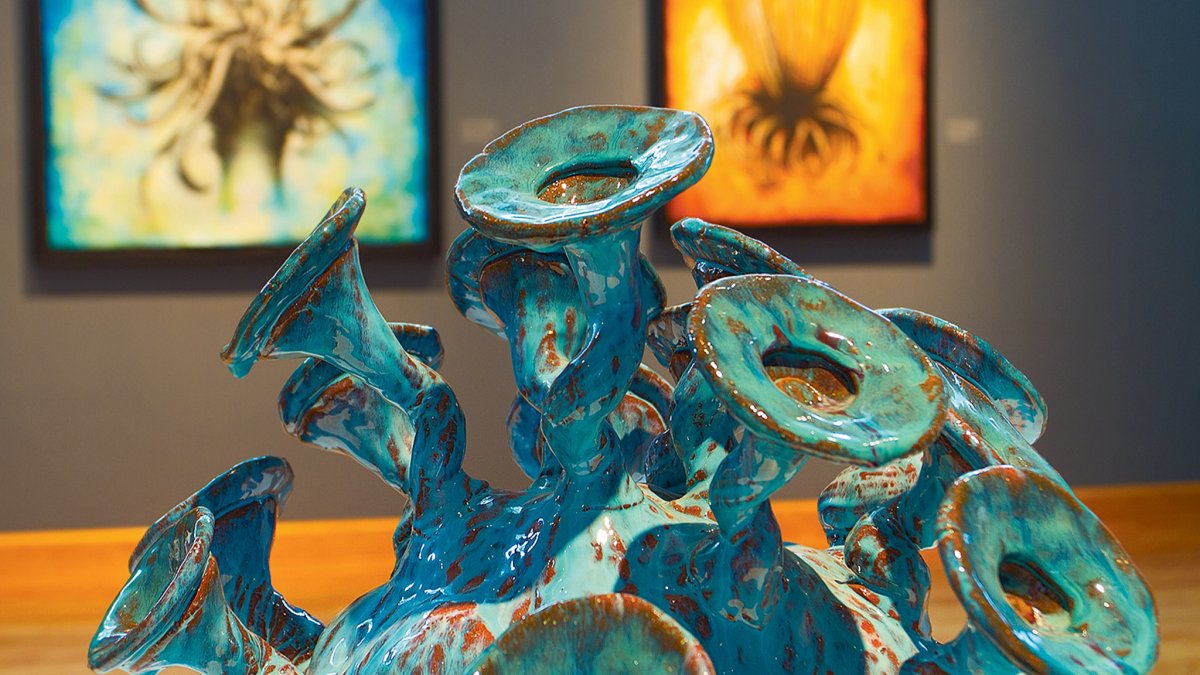 Una obra de arte de cerámica verde azulado y verde en exhibición en el Centro de Bellas Artes de Dunedin con dos pinturas en la pared detrás
