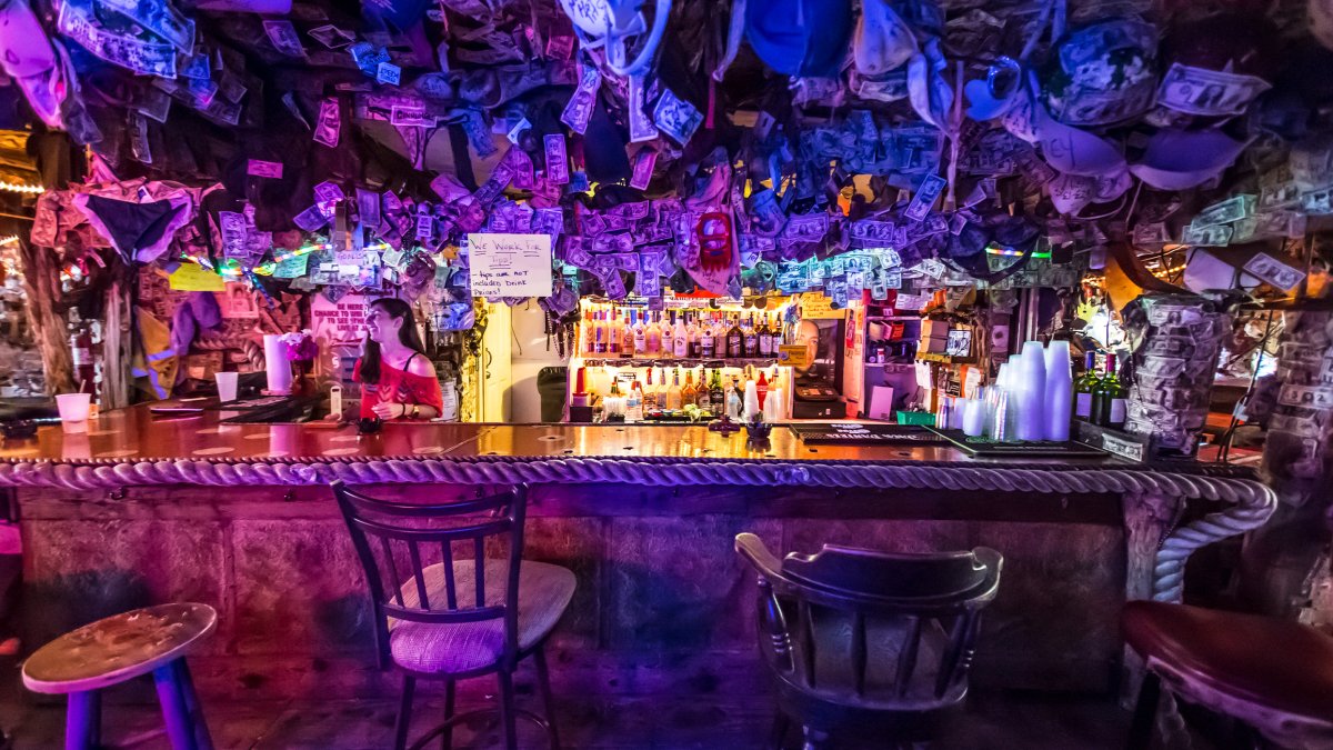 A bartender smiles at a customer at Mahuffer's dive bar; there are dollar bills hanging from the ceiling and purple lights