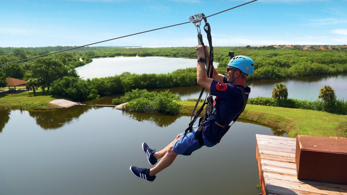 um homem faz tirolesa acima da Mobbly Bayou Wilderness Preserve em Oldsmar; um lago e árvores podem ser vistos abaixo dele