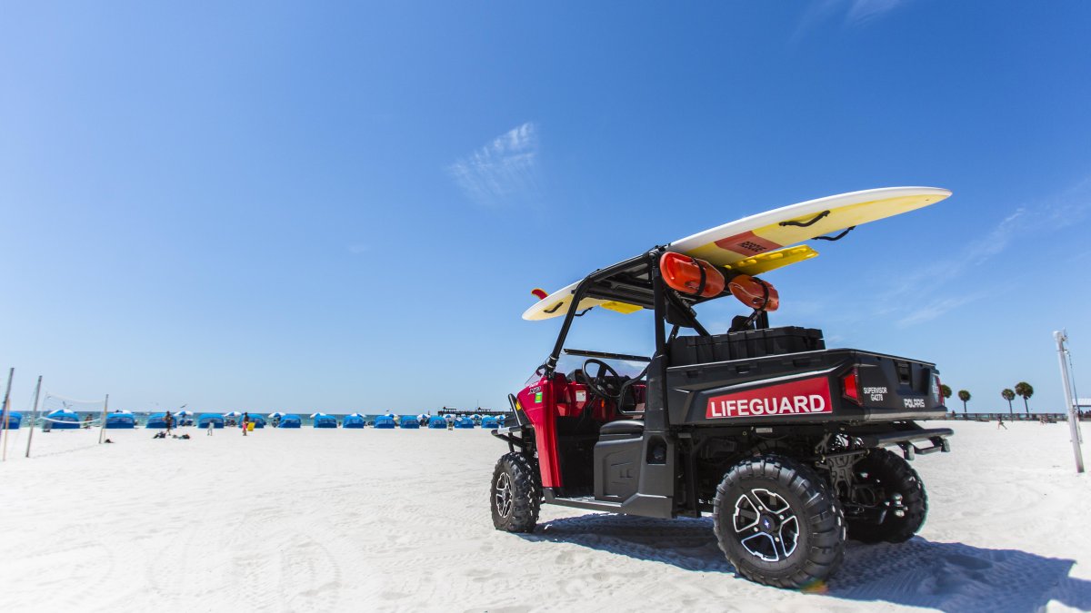 Um buggy salva-vidas estacionado na praia com uma prancha de surf em cima. Cabanas podem ser vistas à distância.