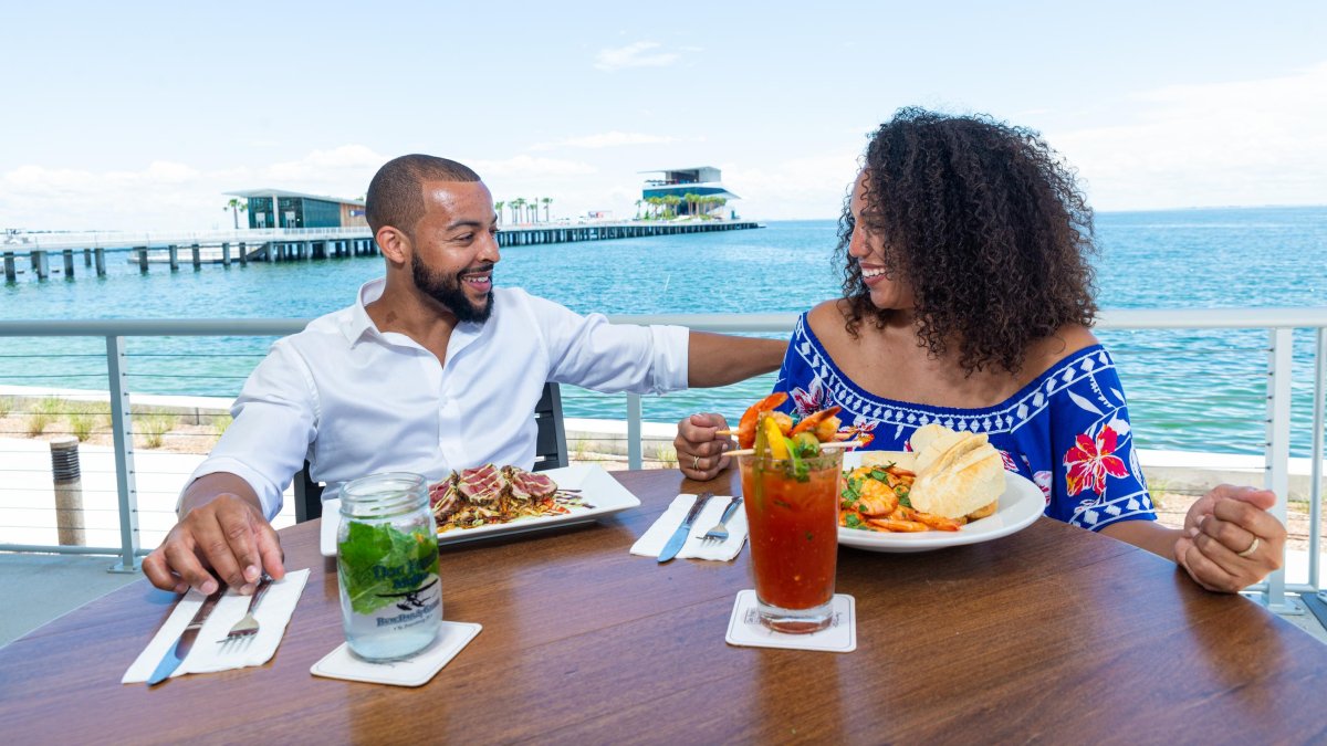 Una pareja cena y disfruta de cócteles en Doc Ford's Rum Bar & Grille con el muelle de St. Pete al fondo.