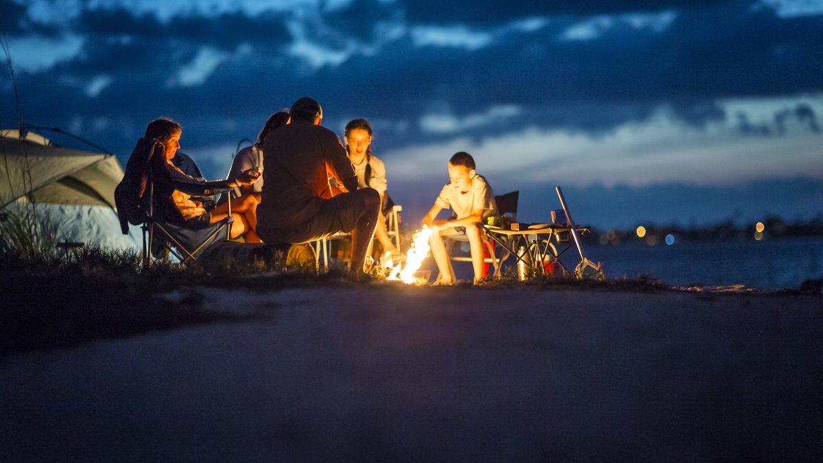 A family circles a bon fire on the beach with a tent in the background