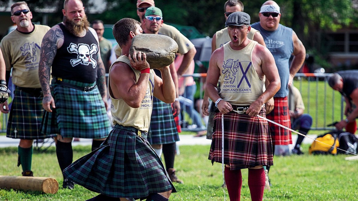 A man dressed in a Scottish outfit trying to throw a massive rock while other men dressed similarly watch.