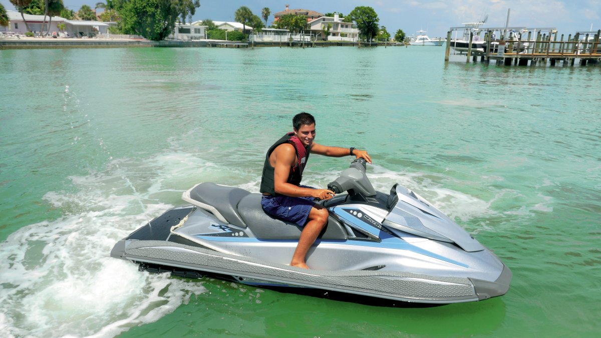 A young man riding a jet ski. Houses and a pier can be seen at a distance.