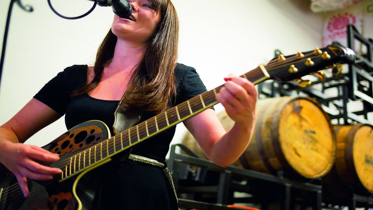 A lady playing a black guitar with beer barrels in the background.