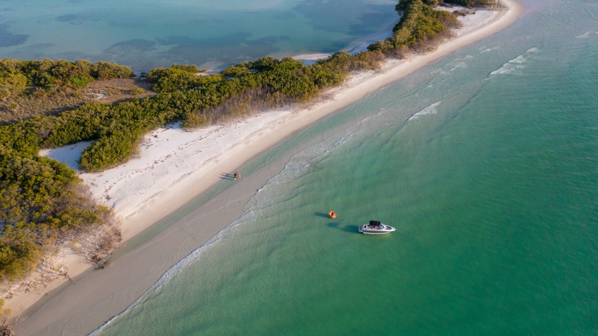 Aerial view of a boat surrounded by green waters and family walking on the nearby pristine beach