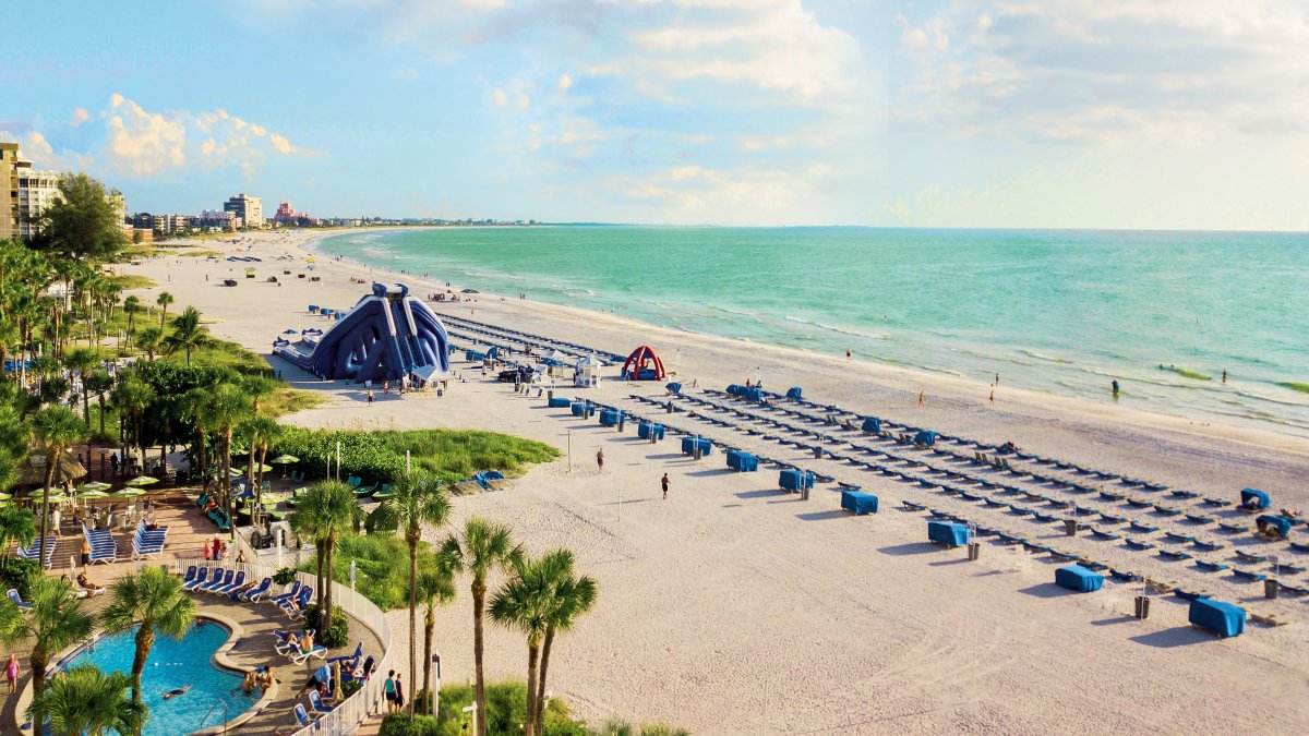 Überblick über den Strandpool des Tradewinds Resort mit blauen Cabanas, einer Wasserrutsche und aquamarinem Wasser.