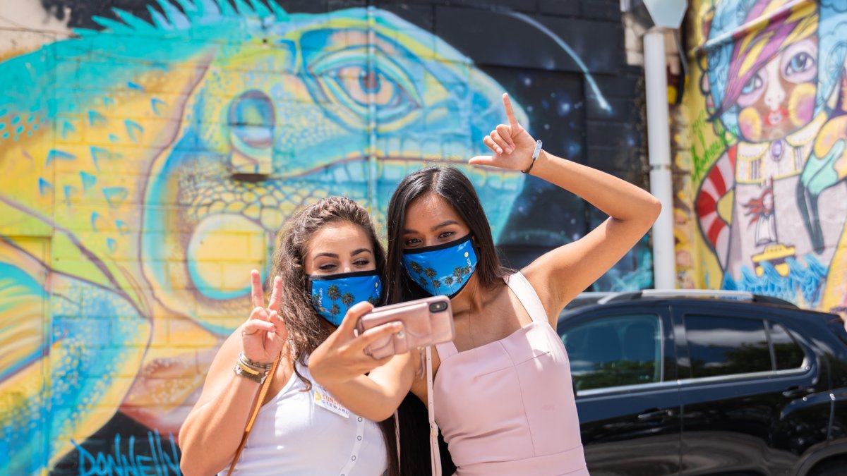 Dos mujeres con máscaras tomándose una selfie frente al mural Lizard King de Derek Donnelly.