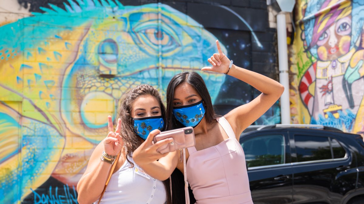 Dos mujeres con máscaras tomándose una selfie frente al mural del Rey Lagarto de Derek Donnelly.