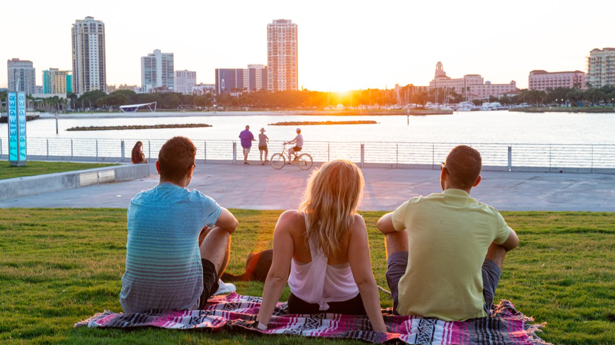 Un grupo de amigos sentados en el campo de hierba cerca del St. Pete Pier viendo la puesta de sol detrás de los edificios