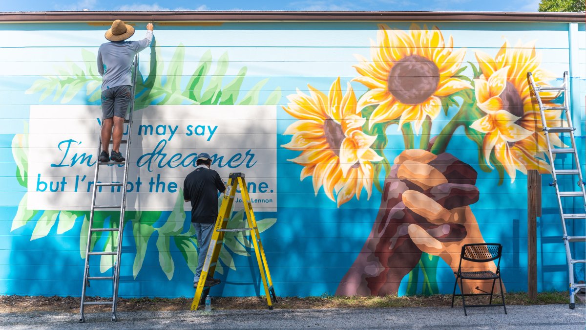 Two people painting hands holding sunflowers and a quote by John Lennon in a mural in St. Pete