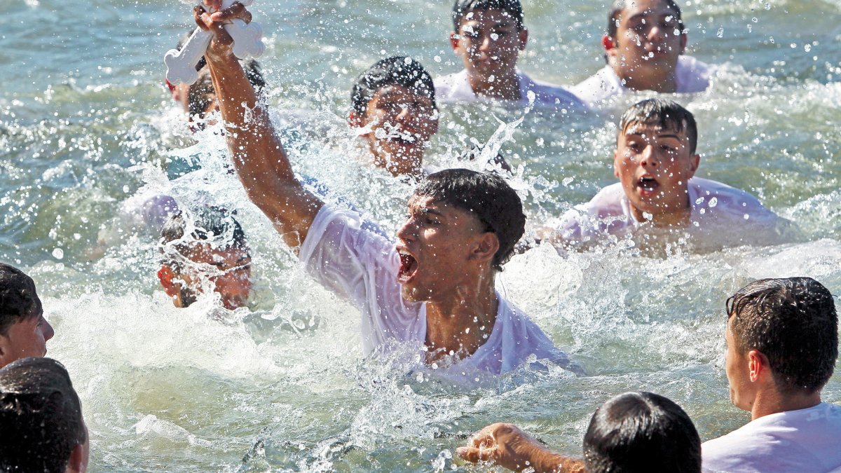 A group of young men in water. One man in the center is holding a cross.