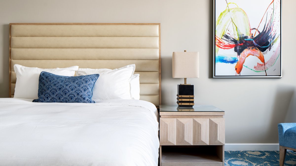 Showing the inside of a hotel room, queen size bed, blue and white carpet, nightstand and abstract painting on the wall.