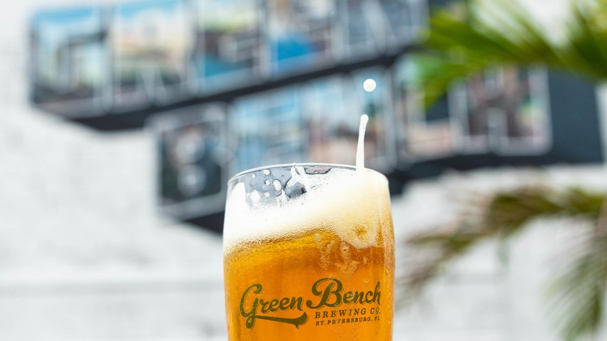 A beer is held up in front of the a Green Bench art mural