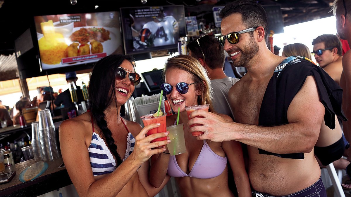 Two Girls and a Guy with shirt over his shoulder having drinks at a beach bar giving cheers.