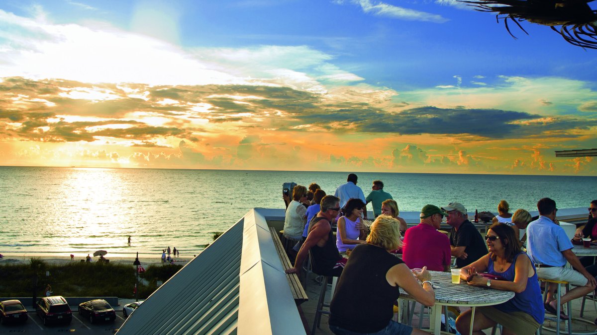 Roof top bar with people enjoying drinks and an ocean view with a sunset in background.