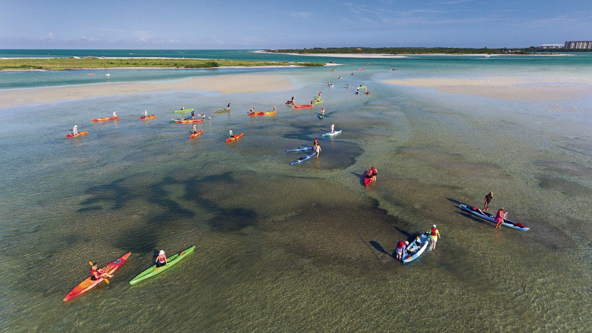 A large group of Kayakers in crystal clear water surrounded by islands. Some are paddling and some are walking in shallow water around there Kayak. 