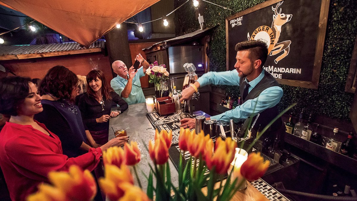 Grupo de pessoas sentadas em um bar com um barman servindo bebidas. O primeiro plano tem um vaso cheio de lírios