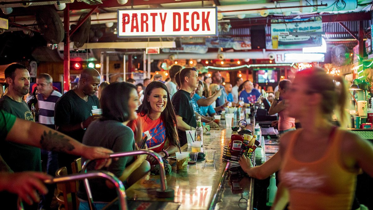 Lots of people sitting and standing at a bar watching sports and drinking, big sign at the top of the images that says "Party Deck"