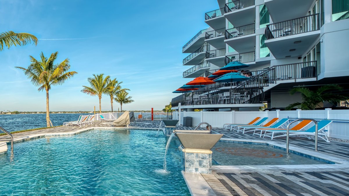 Poolside view of the Hotel, with intercostal waters to the left, pool in the middle and hotel to the right lined with palm trees.