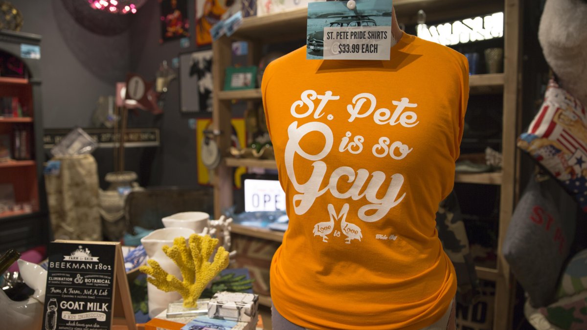 Eine Nahaufnahme eines orangefarbenen T-Shirts mit der Aufschrift „St. Pete is so Gay“. Weitere Waren und Souvenirs sind ausgestellt.