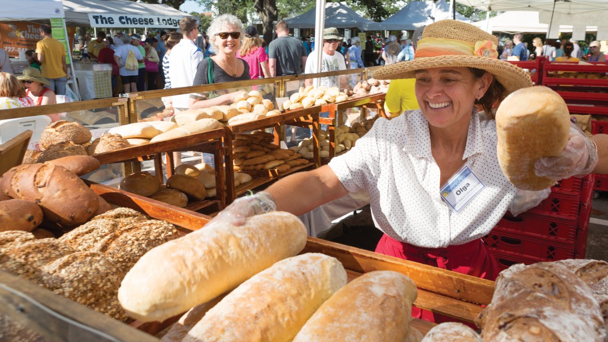 A lady wearing a hat, selling bread with a big smile. Many types of breads are on display.