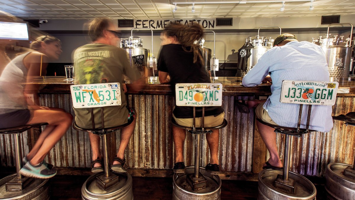 Few people seating at the bar. Bar stools are made of metal barrel and old Floridian license plates.