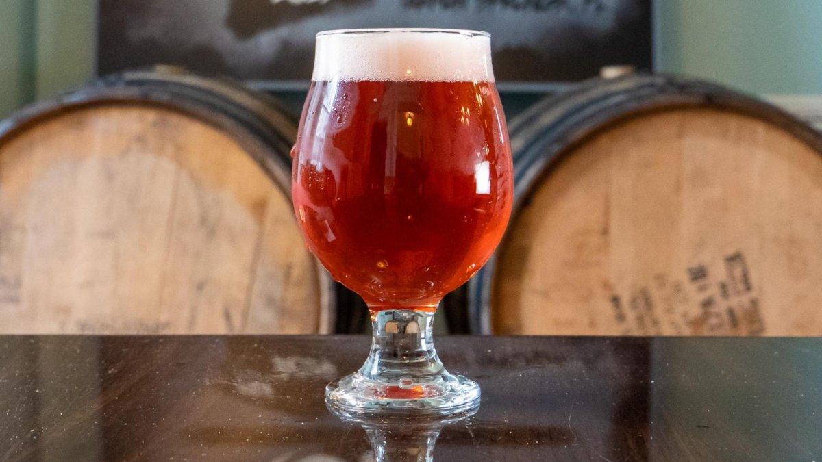 A round glass with red ale in front of wooden barrels.