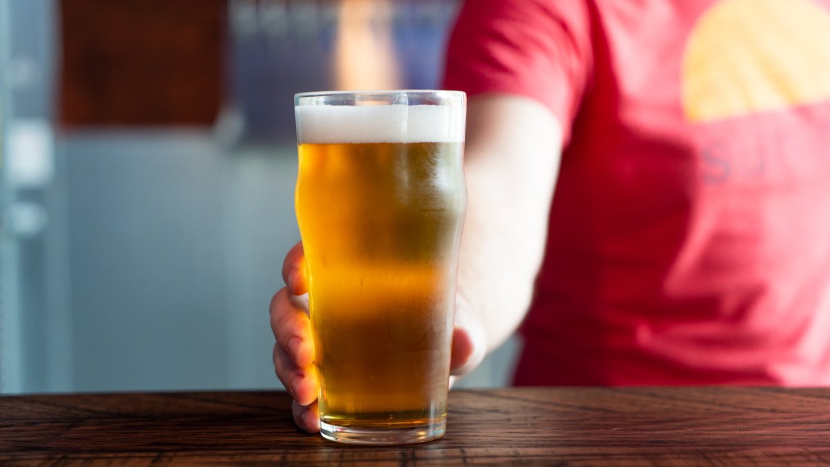 A person wearing a red shirt holds a glass of beer on top of a wooden table.