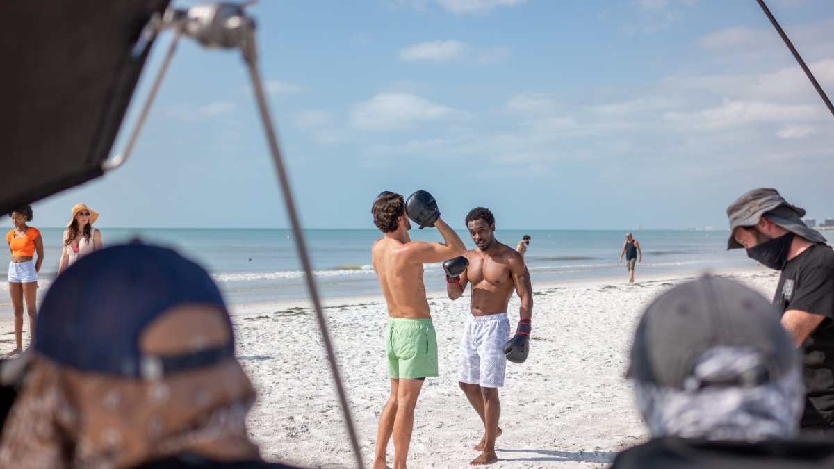 Two men box on the beach in the background with video equipment in the foreground.