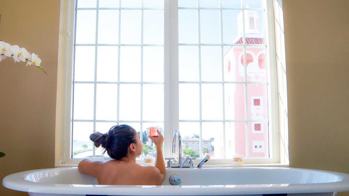 A woman in a bath tub holds a pink champagne glass while looking out the window at the Don Cesar at Spa Oceana