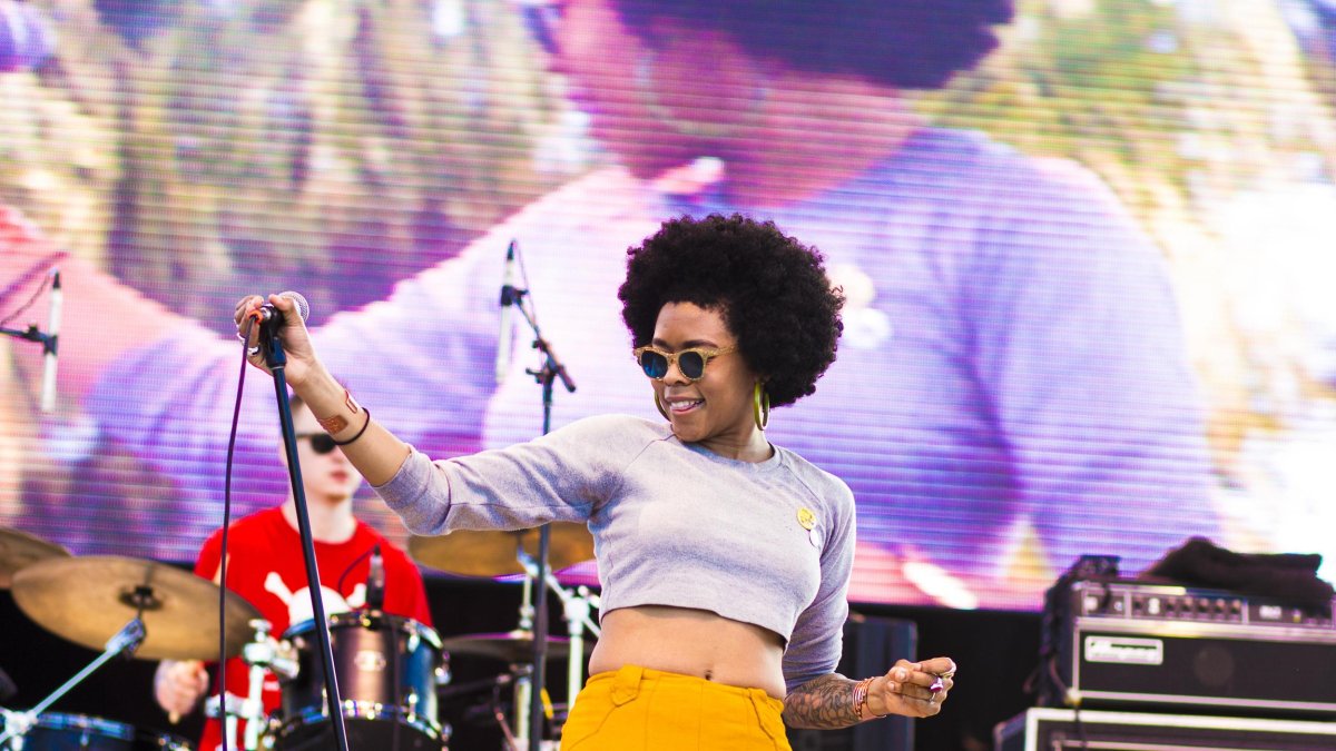 Woman stands on stage holding a microphone stand while a drummer plays behind her.