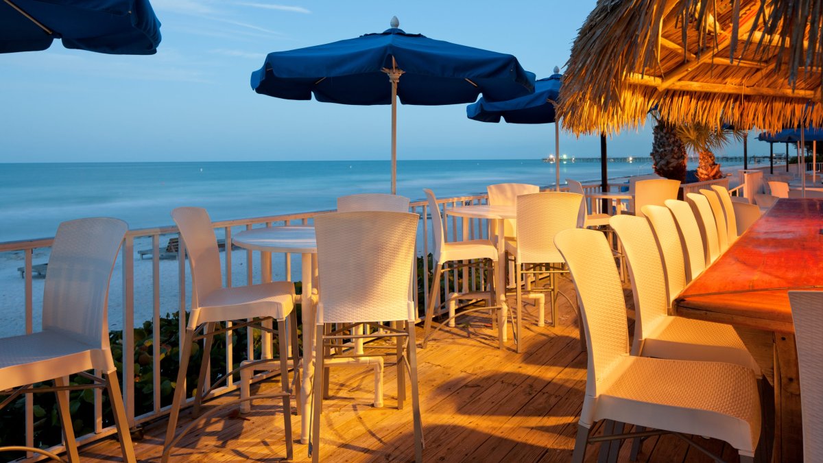 Eine Terrasse am Wasser mit Tischen und Sonnenschirmen in der Abenddämmerung im Mangos DoubleTree Beach Resort by Hilton Tiki Bar