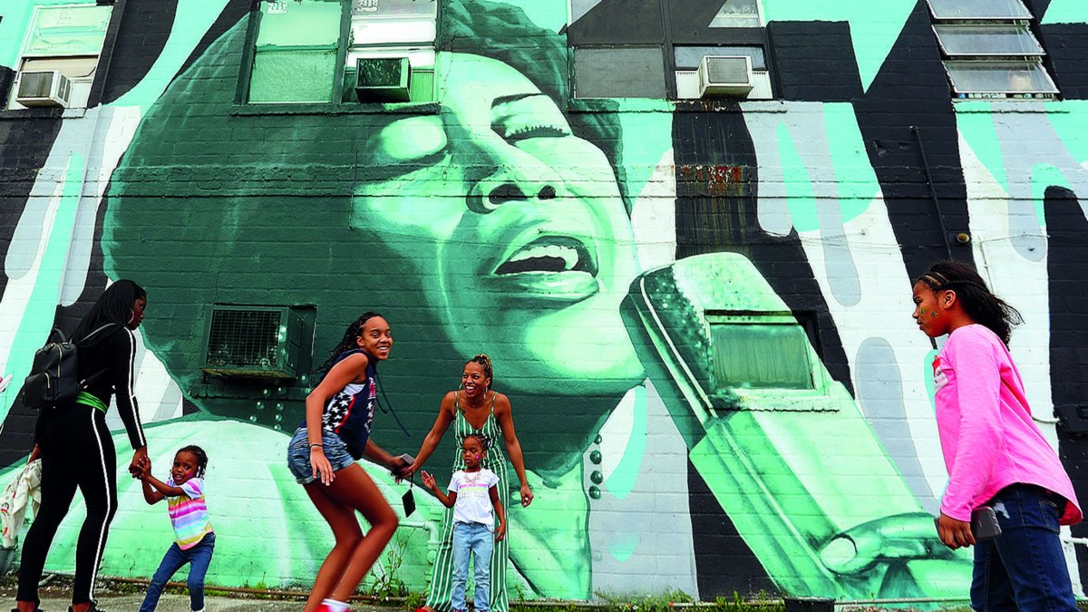 An emotional green and black mural called "Jazz" depicting African American singer Ella Fitzgerald by ZuluPainter; children can be seen playing in front of the mural