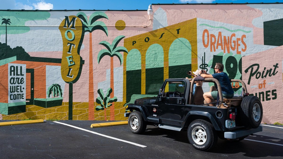 A man and woman in a black jeep park in front of a vintage-themed mural with signs for "motel," "oranges" and "all are welcome here"