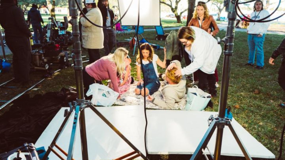 A family sits on a blanket on the grass outside while people setup cameras, hair, and makeup.