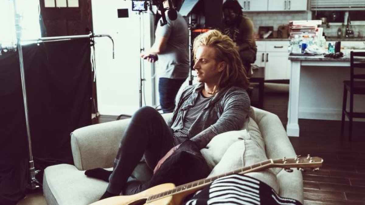 The singer sits on a couch next to a guitar with a filming crew in the background.