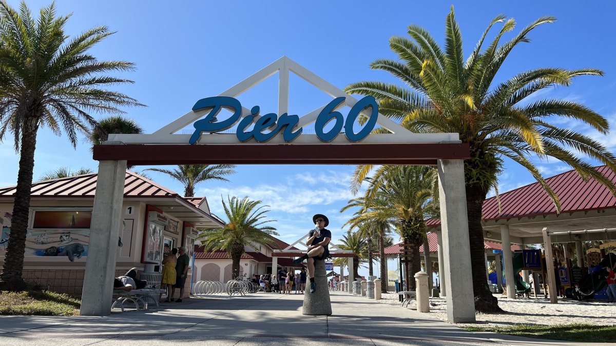 Man sitting under the Pier 60 sign at Clearwater Beach drinking water during the day. Featuring palm trees and sunny skies.